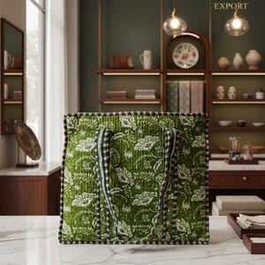May include: Green and white floral patterned tote bag with striped handles. The bag features a quilted design and a black and white trim. The bag is on a white marble countertop. The background includes shelves with decorative items.