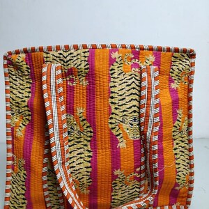 May include: A quilted tote bag with a vibrant tiger print. The bag features a repeating pattern of tigers against a backdrop of vertical orange and pink stripes. The handles and trim are accented with white and orange stripes.