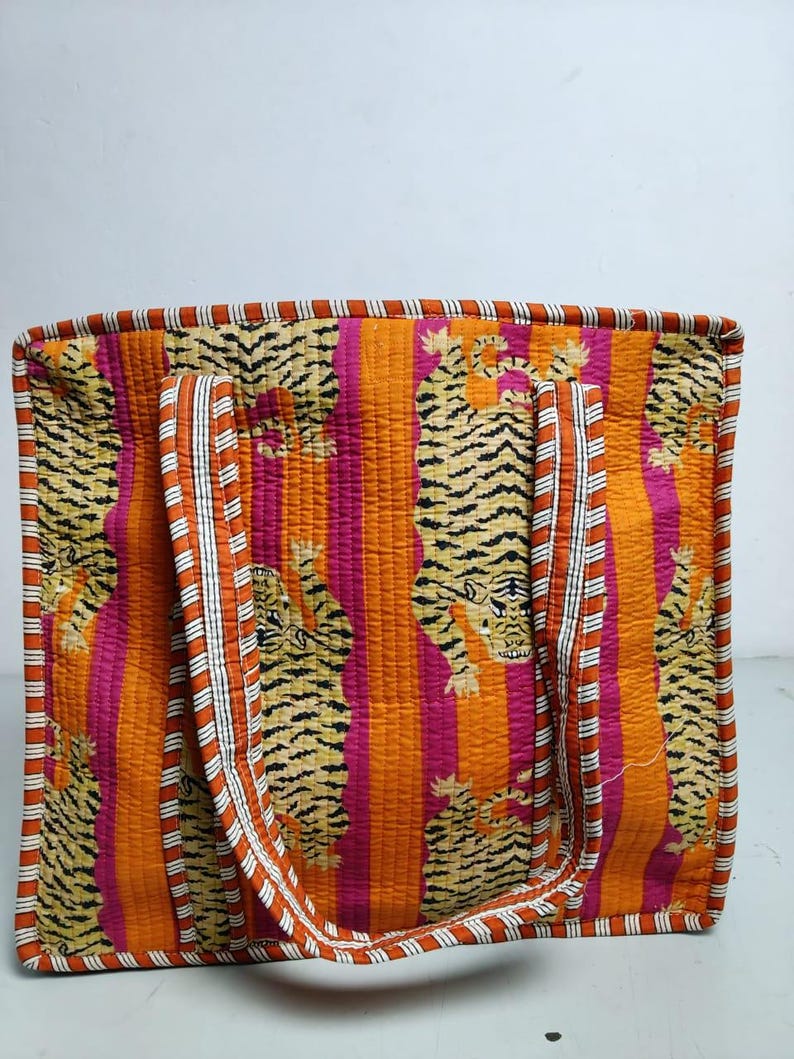 May include: A quilted tote bag with a vibrant tiger print. The bag features a repeating pattern of tigers against a backdrop of vertical orange and pink stripes. The handles and trim are accented with white and orange stripes.