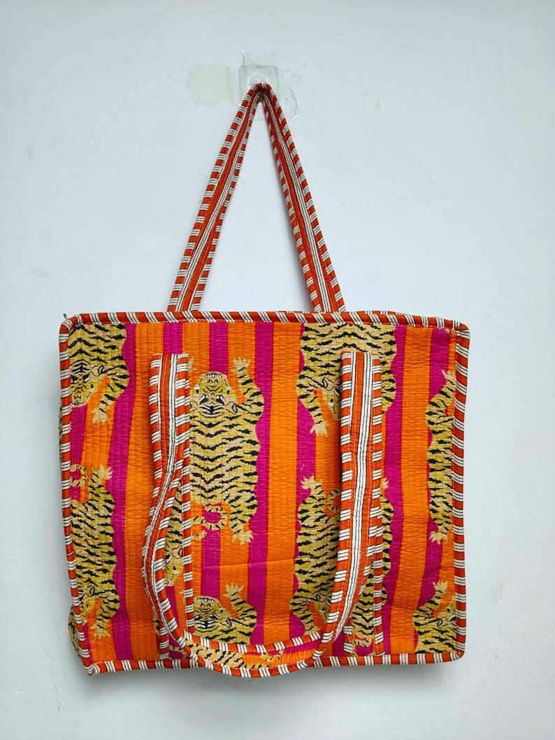 May include: A vibrant tote bag with a repeating tiger pattern on a striped orange and pink background. The bag has matching orange and white striped handles and trim. The bag is made of a quilted fabric.
