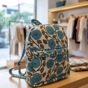 May include: A blue and white floral patterned backpack. The bag features a front pocket and adjustable straps. The backpack is made from a textured fabric and is displayed on a wooden surface. Clothing racks and shelves are visible in the background.