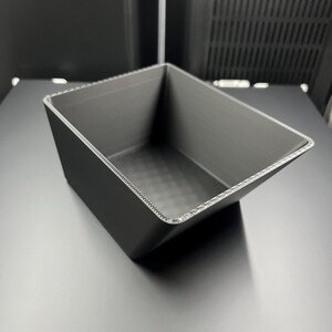 Pode incluir: Um recipiente retangular cinza escuro com laterais anguladas. O recipiente tem uma superfície texturizada e uma borda decorativa. O objeto é provavelmente impresso em 3D e está sobre uma superfície plana.