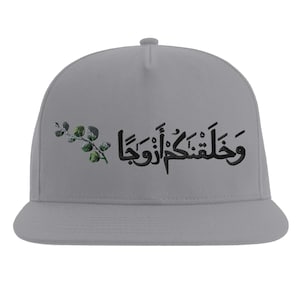 Può includere: Cappellino da baseball grigio chiaro con visiera piatta. Il cappellino presenta una scritta araba nera e un piccolo motivo floreale verde e grigio sul lato sinistro. Il testo è centrato sul davanti.
