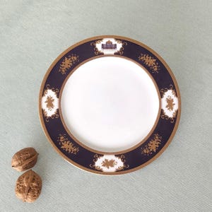 Könnte beinhalten: Ein weißer Keramikteller mit einem marineblauen und goldenen Zierrand. Der Rand weist verzierte goldene Blumen- und Architekturmuster auf. Zwei Walnüsse liegen neben dem Teller auf einer hellgrünen Oberfläche.