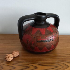 May include: A ceramic vase with a black handle and rim. The body of the vase is brown with red and brown geometric patterns. Two walnuts sit on the wooden surface in front of the vase.