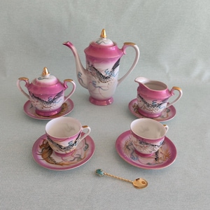Op de afbeelding: Een vintage porseleinen theeservies met een roze en wit ontwerp, bestaande uit een theepot, suikerpot, melkkan, twee kopjes, schotels en een gouden lepel. De set heeft een drakenmotief en gouden accenten.