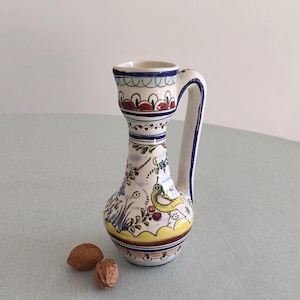 Könnte beinhalten: Eine dekorative Keramikkrug mit weißem Hintergrund, mit einem bunten Vogel- und Blumendesign. Der Krug hat einen Griff und eine kleine Öffnung. Zwei Walnüsse liegen vor dem Krug.