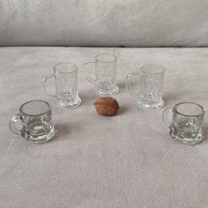 May include: Four miniature clear glass beer mugs with sturdy handles and a textured base design, arranged in a semi-circle around a single brown walnut shell. Each small mug features a thick rim and a clear, transparent glass construction.