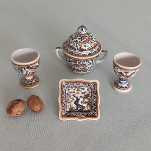 Może przedstawiać: Zestaw ceramiczny składający się z cukiernicy z pokrywką i dwoma uchwytami, dwóch kieliszków do jajek na nóżce oraz małego kwadratowego talerzyka, ozdobionych ręcznie malowanymi motywami kwiatowymi i zwierzęcymi w brązie, błękicie i zieleni, z dwiema orzechami włoskimi.