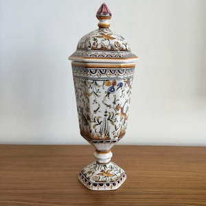 Może przedstawiać: Wysoki, sześciokątny ceramiczny pojemnik z pokrywką, ozdobiony ręcznie malowanymi motywami ptaków, jeleni i kwiatów w odcieniach brązu, błękitu i żółci. Naczynie stoi na podstawie w formie cokołu i posiada dopasowaną pokrywkę ze szpicem.