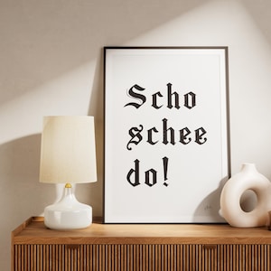 Puede incluir: Una impresión enmarcada con las palabras "Scho schee do!" en letras góticas negras. Una lámpara blanca con una pantalla beige, un jarrón blanco y un pequeño jarrón marrón con flores secas están sobre un mueble de madera.