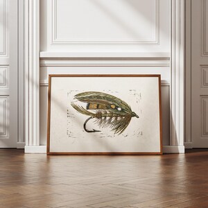 Könnte beinhalten: Ein gerahmter Druck einer Angel-Fliege. Das Kunstwerk zeigt eine detaillierte Illustration einer Fliege mit braunen, grünen und beigen Federn, einem schwarzen Haken und weißen Akzenten. Der Druck befindet sich in einem braunen Holzrahmen, der an einer weißen Wand lehnt.