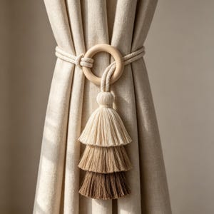 May include: A beige curtain tieback with a wooden ring and a layered tassel in shades of beige and brown. The tieback is made of rope and is wrapped around the curtain. The tassel hangs down, adding a decorative element to the window treatment.
