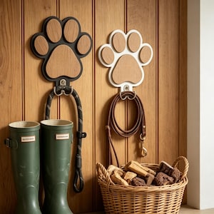 May include: Two paw-shaped wall hooks, one black and one white, with wooden paw prints. Below, a black leash hangs from the black hook, and a brown leash from the white hook. Green rain boots and a wicker basket filled with dog treats complete the scene.
