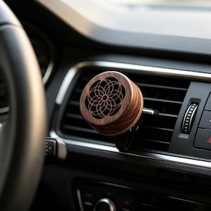 May include: A brown wooden car air freshener with a geometric floral pattern. The air freshener is attached to a car vent. The car's interior is black.