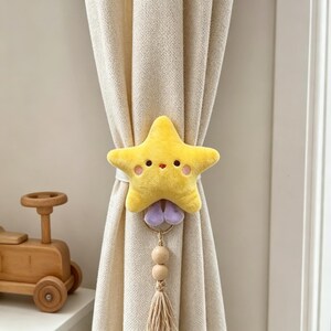 May include: A yellow plush star curtain tieback with a smiling face and pink cheeks. The tieback has a purple accent, wooden beads, and a beige tassel. The curtain is a light beige color.