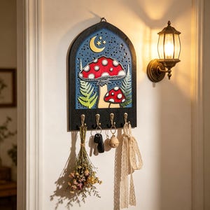 May include: A decorative key holder with a whimsical mushroom design. The key holder features a red and white mushroom, a blue night sky with a crescent moon and stars, and four brass hooks. A small bouquet of dried flowers and keys hang from the hooks.