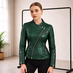Könnte beinhalten: Eine dunkelgrüne Lederjacke mit asymmetrischem, silberfarbenem Reißverschluss, Stehkragen mit Druckknopf, gesteppten Schulterpartien, Reißverschlusstaschen an Brust und Ärmeln sowie verstellbaren seitlichen Schnallen an der Taille.