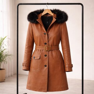 May include: A brown leather coat with a black fur-trimmed hood, belted waist, and button-down front. The coat has two front pockets and is hanging on a black clothing rack.