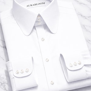 Puede incluir: Una camisa blanca impecable con cuello italiano y puños abotonados. La camisa está cuidadosamente doblada, con el nombre de la marca "AURABLOOM" visible en el cuello. Diseño clásico, ideal para ocasiones formales.