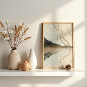 May include: A framed landscape art print with a light wood frame. The artwork depicts a mountain scene reflected in water, with beige and brown tones. Decorative vases and a pinecone sit on a white shelf.