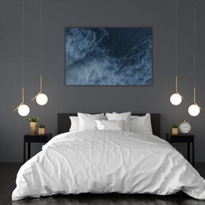 Op de afbeelding: Een moderne slaapkamer met een groot bed bedekt met wit beddengoed. Boven het bed hangt een blauw abstract kunstwerk. Twee zwarte nachtkastjes flankeren het bed, elk met een kleine plant en een goudkleurige lamp. Gouden hanglampen hangen aan het plafond.