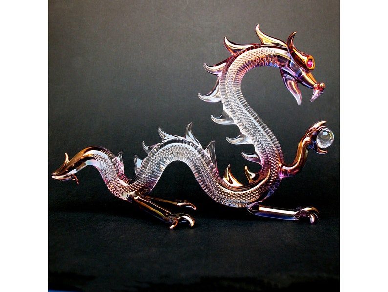 May include: A glass dragon figurine with iridescent pink and gold coloring. The dragon is curled in a defensive position with its claws extended and a small glass orb in its right paw.