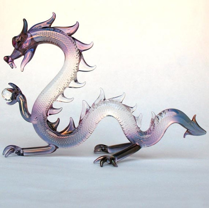May include: A glass dragon figurine with iridescent purple and white coloring. The dragon is holding a clear glass orb in its front paw.