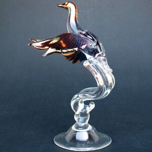 May include: A glass sculpture of a bird with iridescent purple and brown wings, perched on a clear glass stand with a swirling design.