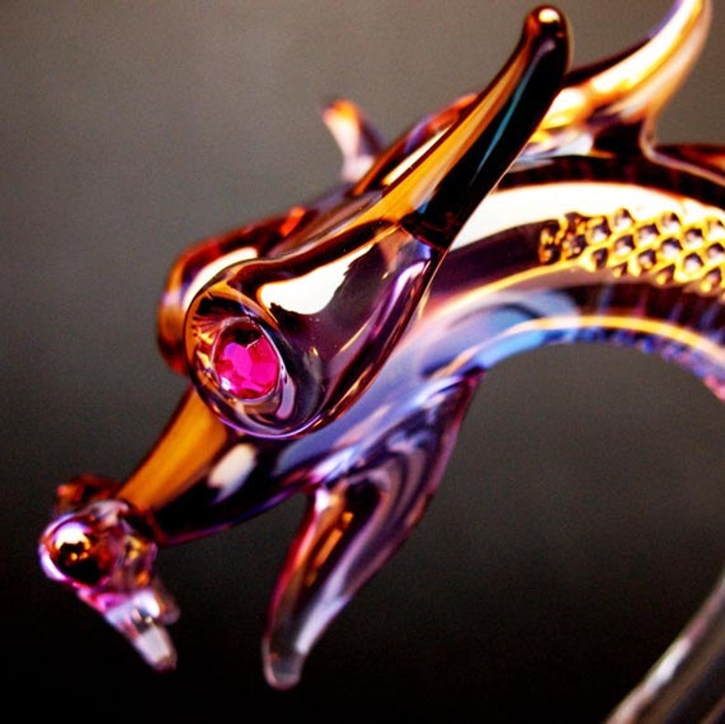 May include: A glass dragon figurine with iridescent purple and orange hues. The dragon has a pink gem for an eye.