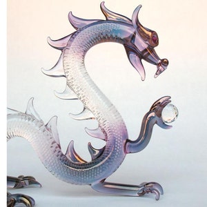 May include: A glass dragon figurine with a purple and white gradient. The dragon is holding a clear glass orb in its paw.