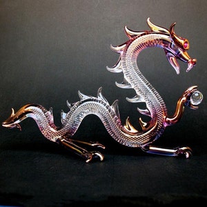 May include: A glass dragon figurine with iridescent pink and white coloring. The dragon is curled in a defensive position with its head raised and its claws extended. The dragon has a crystal ball in its right claw.