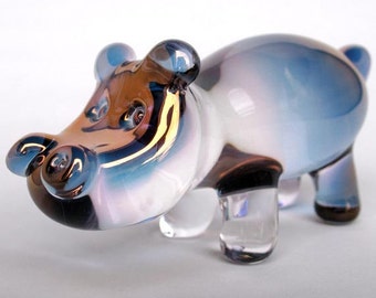 May include: A small glass figurine of a hippopotamus with a blue, white, and brown iridescent glaze. The hippo has a rounded body and short legs.