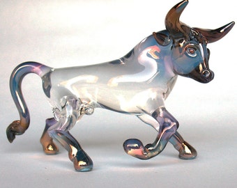 May include: A clear glass bull figurine with iridescent accents. The bull is standing with its head turned to the side and its horns pointing up. The figurine is a decorative piece for a home or office.