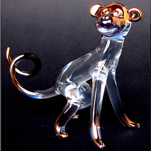 May include: A clear glass monkey figurine with a curled tail. The monkey's head and limbs are accented with a metallic gold finish.