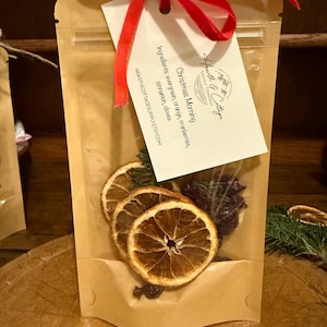 May include: A clear cellophane bag filled with dried orange slices, cranberries, and evergreen sprigs. A tag tied with a red ribbon reads "Christmas Morning" with ingredient details. The bag is on a wooden surface.