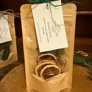 May include: A clear cellophane bag filled with dried orange slices, evergreen sprigs, and a tag that reads "Christmas Tree Farm." The tag lists ingredients: evergreen, orange, cinnamon, cloves, and anise. The bag is tied with green twine.
