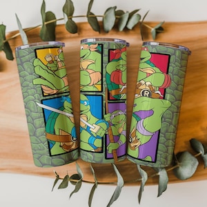 Puede incluir: Tres vasos de acero inoxidable con las Tortugas Ninja. Cada vaso tiene un patrón de escamas verdes y una ilustración de personaje diferente en un diseño enmarcado. Las ilustraciones incluyen a las tortugas en poses de acción con sus armas características.