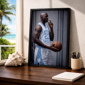Puede incluir: Impresi&oacute;n enmarcada de un jugador de baloncesto con una camiseta azul claro y blanca con el n&uacute;mero 23. El jugador sostiene un bal&oacute;n de baloncesto y un cigarro. La impresi&oacute;n est&aacute; sobre una superficie de madera cerca de una ventana.