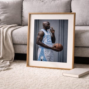 Puede incluir: Impresi&oacute;n enmarcada de un jugador de baloncesto con una camiseta azul claro y blanca con el n&uacute;mero 23. El jugador sostiene una pelota de baloncesto y un cigarro. La impresi&oacute;n est&aacute; en un marco de madera, sobre una alfombra blanca.