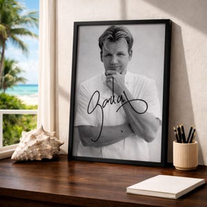 May include: Black and white framed print featuring a portrait of a chef in a white uniform, with a signature in black ink. The print is displayed on a wooden surface, next to a seashell and a pen holder. The background shows a beach scene.