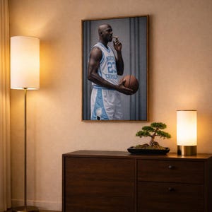 Puede incluir: Una impresi&oacute;n enmarcada de un jugador de baloncesto con una camiseta azul claro con el n&uacute;mero 23, sosteniendo una pelota de baloncesto y un cigarro. La impresi&oacute;n cuelga sobre un mueble de madera oscura, con un bons&aacute;i y una l&aacute;mpara.