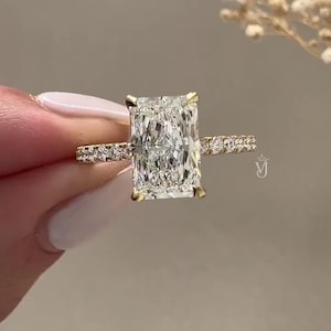 Może przedstawiać: Złoty pierścionek zaręczynowy z diamentem o szlifie prostokątnym. Główny diament jest utrzymywany przez cztery pazury. Opaska jest ozdobiona mniejszymi diamentami. Po prawej stronie pierścionka widoczny jest symbol.