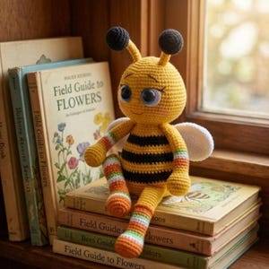 May include: A crocheted bee doll with a yellow and black striped body, large blue eyes, and two antennae topped with black spheres. The doll has striped arms and legs in orange, white, and green, and small white wings attached to its back.
