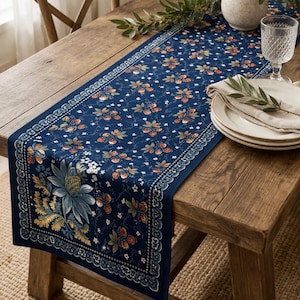 May include: A navy blue table runner with a floral pattern featuring orange and white flowers and green leaves. The runner is placed on a wooden table, with a decorative border and a large floral design at one end. A glass and plates are on the table.