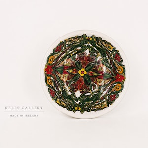 May include: A white ceramic plate with a detailed floral design in red, green, yellow, and brown. The plate features a central flower and intricate patterns. Text on the plate reads "KELLS GALLERY" and "MADE IN IRELAND".