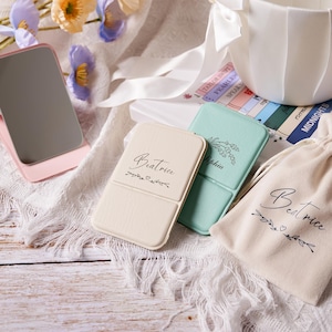 Op de afbeelding: Een verzameling gepersonaliseerde compacte spiegels en een stoffen zakje. De spiegels zijn in pastelkleuren, met de naam "Beatrice" en bloemmotieven. Eén spiegel is mintgroen met de naam "Sophia". Het zakje heeft ook de naam "Beatrice".