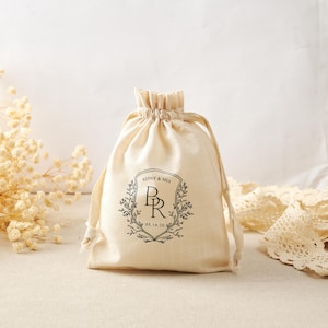 Puede incluir: Una bolsa de tela beige con cordón que presenta un diseño de escudo impreso en negro con las letras B y R, los nombres KENNY & MIA y la fecha 05.14.26, encerrados dentro de una corona botánica decorativa de hojas y ramas.