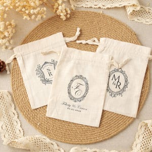 Peut inclure: Trois pochettes à cordon en tissu blanc cassé avec des motifs imprimés noirs. Chaque sac présente un cadre ovale orné unique contenant des initiales et des dates, dont le texte Felicity & Carson 02.07.2026, Salina & Ryan et M & R.