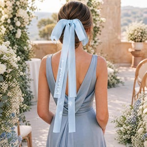 May include: A light blue satin hair ribbon tied in a bow, featuring the name Rosamund printed in white script on one of the long, trailing tails. The ribbon is secured in a hairstyle, complementing a matching light blue sleeveless dress.